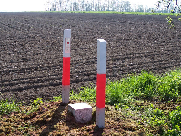 Das Bild zeigt einen Vermessungspunkt im Boden an einem Feld. Dieser wird geschützt durch 2 weißrot markierte Pfäle